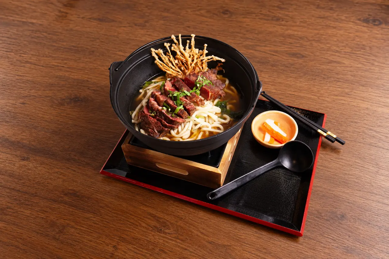 Ribeye Jaew Hon soup with sliced ribeye, udon, herbal broth, and crispy enoki in a black pot.